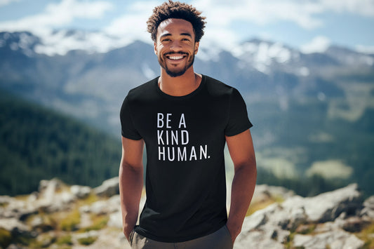 A Black man standing outside on rocky ground wearing a black unisex t-shirt that says, "BE A KIND HUMAN." in a clean, sans-serif, narrow font. There are snow-capped mountains behind him.