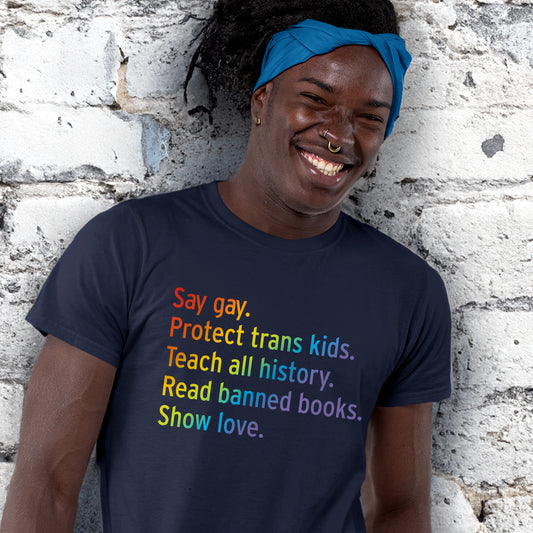 A man of color wearing a Navy Blue t-shirt that says "Say gay. Protect trans kids. Teach all history. Read banned books. Show love."