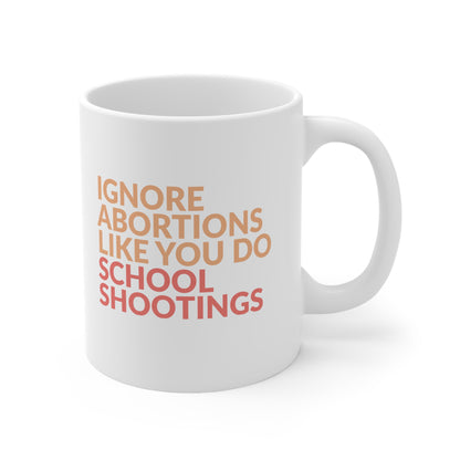 White 11oz ceramic mug that says “Ignore abortions like you do school shootings” in all caps in an orange font. The words “school shootings” are in red. The handle is on the right side in this image.
