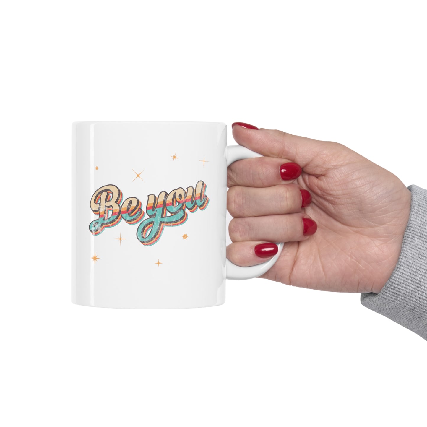 An image of a woman's hand holding an 11oz white ceramic mug that says “Be you” in a vintage-inspired script font with vintage colors and stripes. It’s designed to look as if small parts of the design are coming off of the mug.