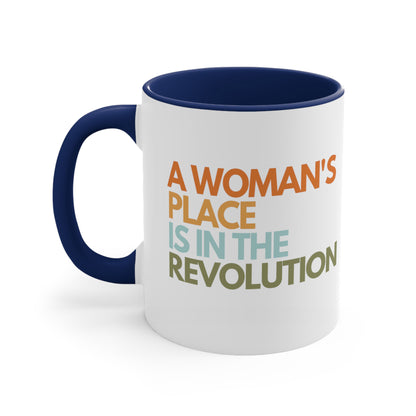 11oz ceramic mug with a navy blue interior and handle that says “A woman’s place is in the revolution” in a round, modern font in all caps and left aligned. Each line is a different color. “A Woman’s” is orange, “place” is yellow-orange, “is in the” is light blue, and “revolution” is military green.