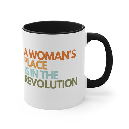 11oz ceramic mug with a black interior and handle that says “A woman’s place is in the revolution” in a round, modern font in all caps and left aligned. Each line is a different color. “A Woman’s” is orange, “place” is yellow-orange, “is in the” is light blue, and “revolution” is military green.