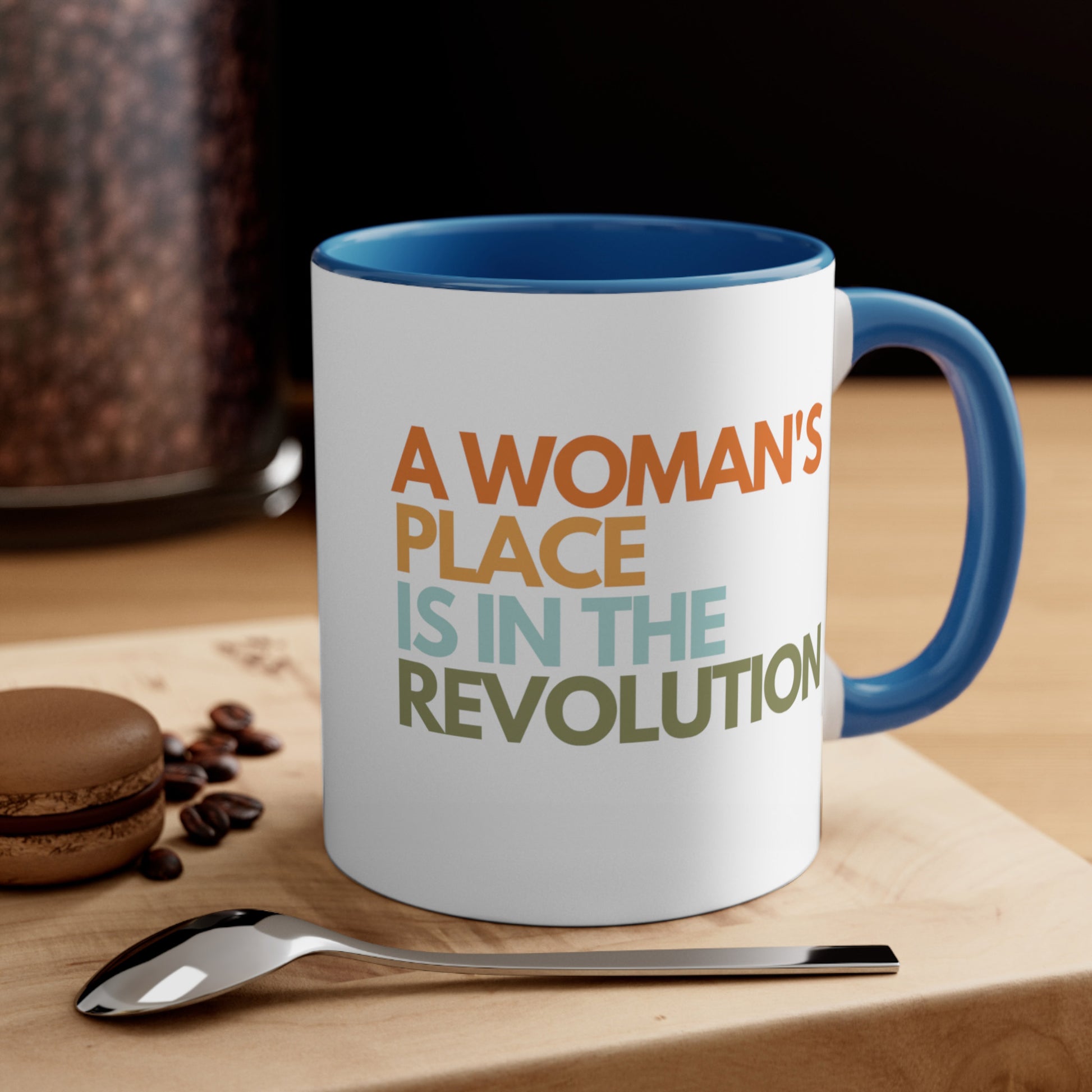 Contextual photo of an 11oz ceramic mug with a blue interior and handle that says “A woman’s place is in the revolution” in a round, modern font in all caps and left aligned. Each line is a different color. “A Woman’s” is orange, “place” is yellow-orange, “is in the” is light blue, and “revolution” is military green. The mug is on a table with a cookie and a spoon.