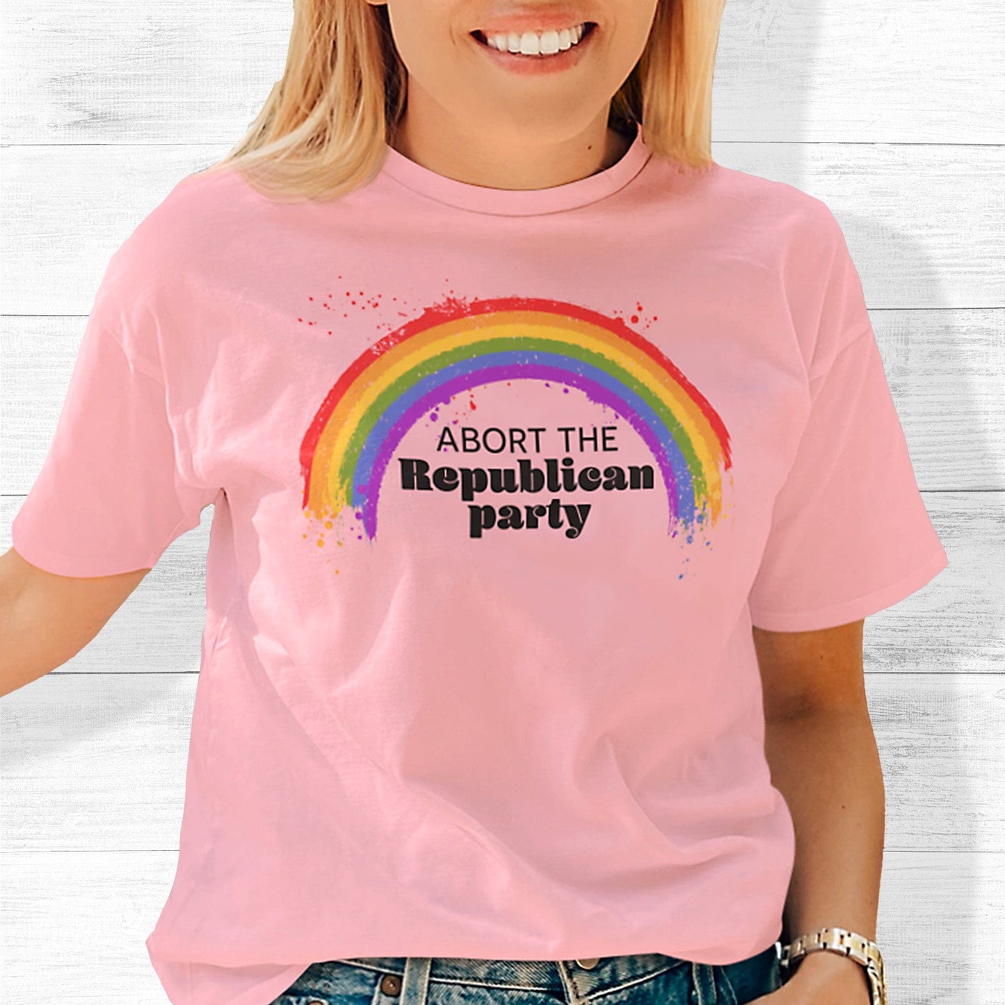 Pink unisex t-shirt with a graphic of a rainbow with small paint splatters around it. Inside the negative space of the rainbow is the text, “Abort the Republican Party.” 