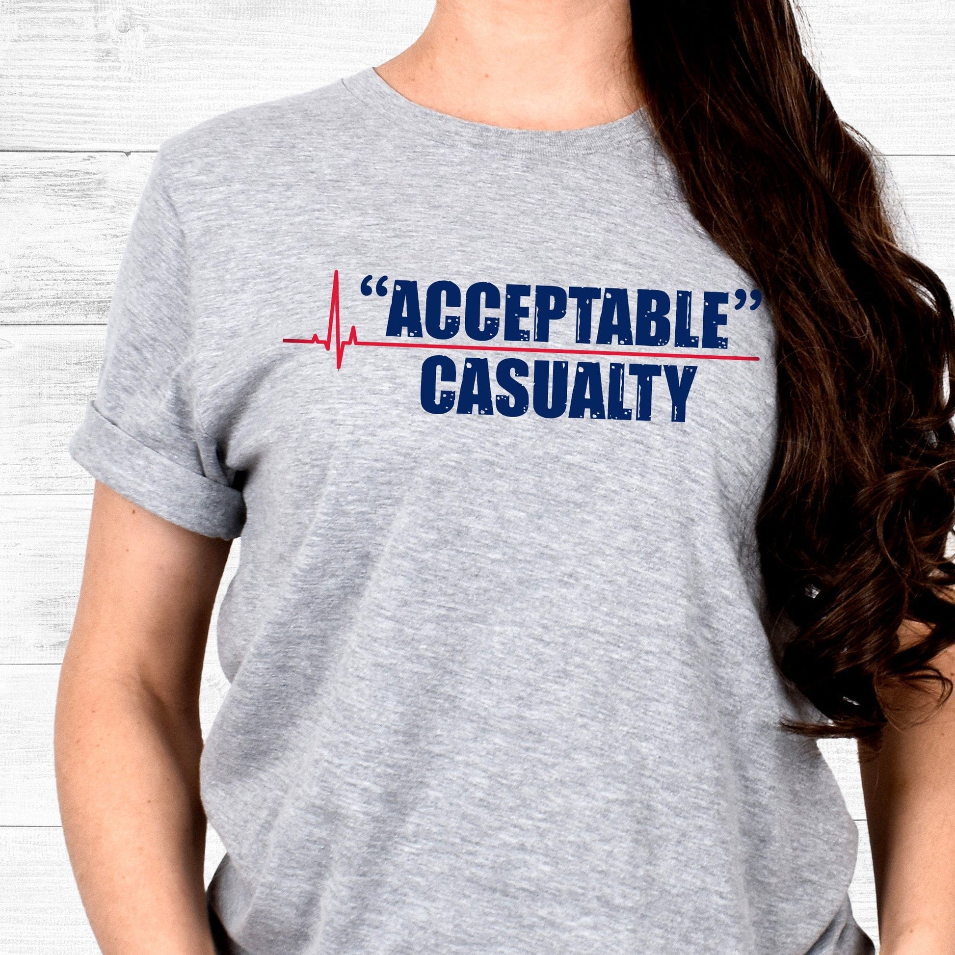 Athletic Heather color unisex t-shirt that says, “Acceptable” Casualty with each word on its own line. There is a red heartbeat graphic to the left of the text with a straight, flat line extending out between the two words.