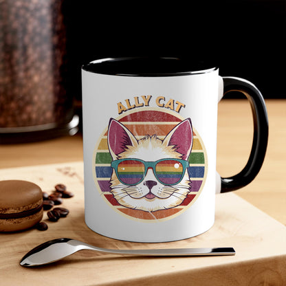 Contextual photo of an 11oz ceramic mug with a black handle and interior with a circular retro-inspired image of a cat wearing rainbow sunglasses with a retro sunset made from parallel lines behind it. The text reads “ALLY CAT” in all caps, arched over the image. The graphic has a retro, worn texture. THe mug is on a table with a spoon and a cookie.