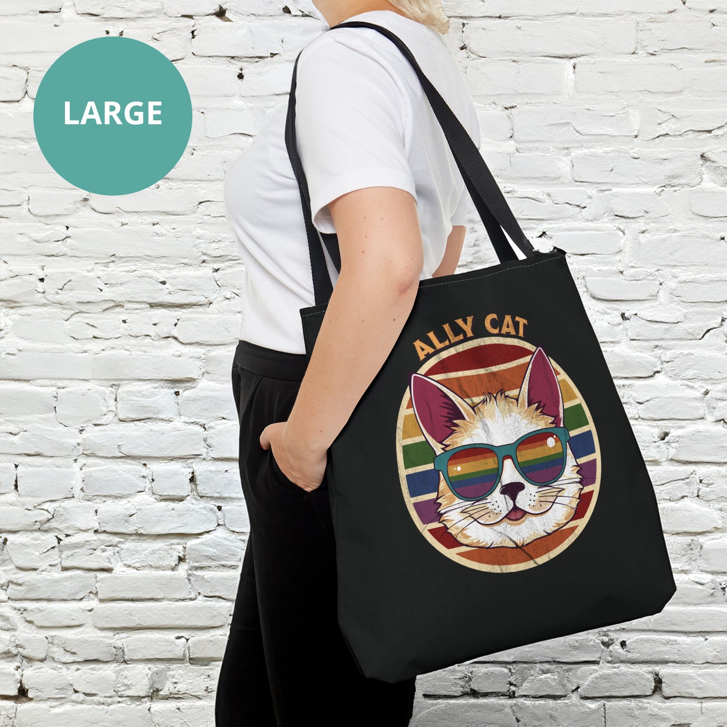 Contextual photo of a black tote bag with a black handle with a circular retro-inspired image of a cat wearing rainbow sunglasses with a retro sunset made from parallel lines behind it. The text reads “ALLY CAT” in all caps, arched over the image. The graphic has a retro, worn texture.