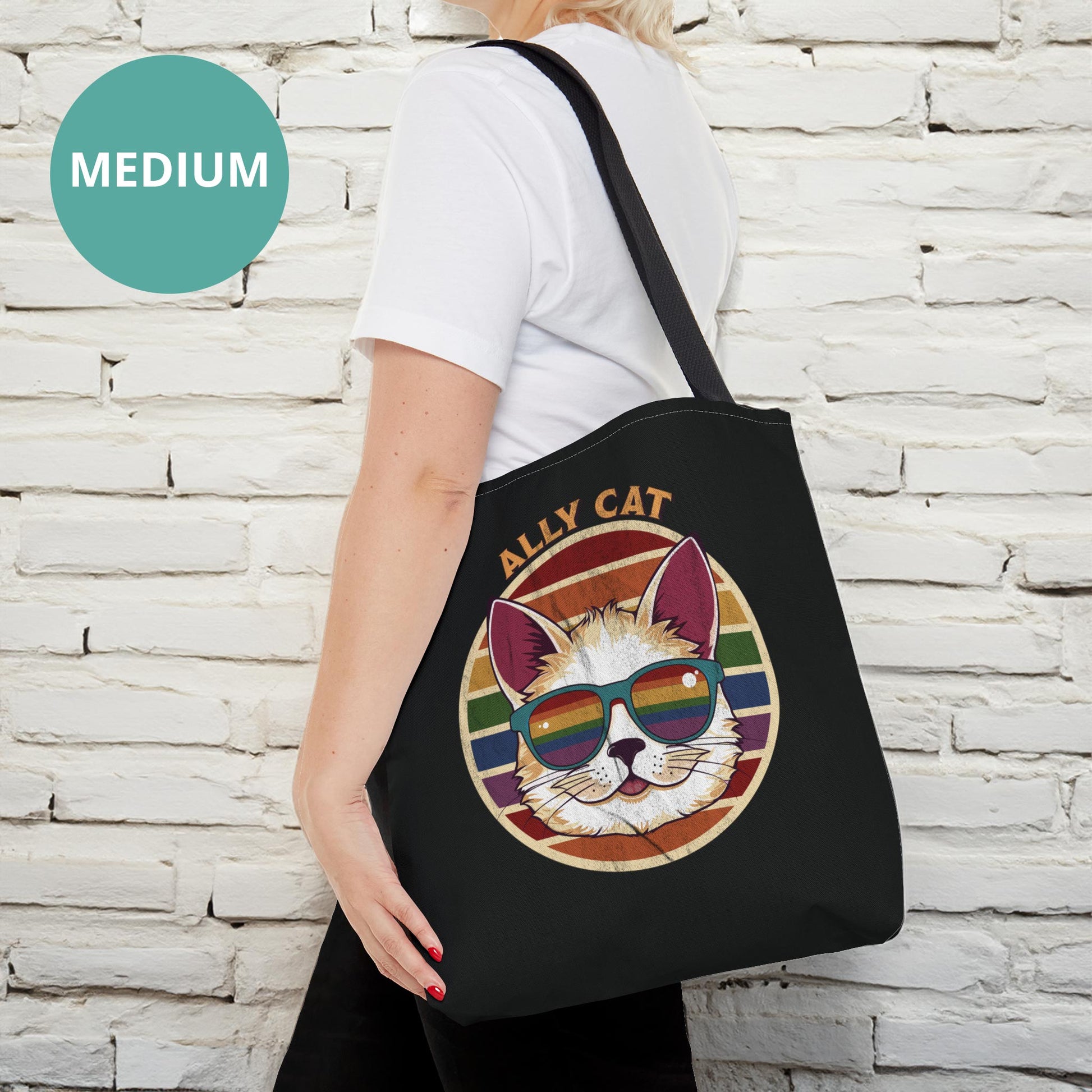 Contextual photo of a black tote bag with a black handle with a circular retro-inspired image of a cat wearing rainbow sunglasses with a retro sunset made from parallel lines behind it. The text reads “ALLY CAT” in all caps, arched over the image. The graphic has a retro, worn texture.