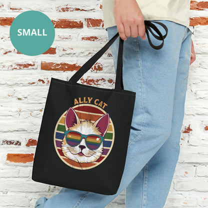 Contextual photo of a black tote bag with a black handle with a circular retro-inspired image of a cat wearing rainbow sunglasses with a retro sunset made from parallel lines behind it. The text reads “ALLY CAT” in all caps, arched over the image. The graphic has a retro, worn texture.