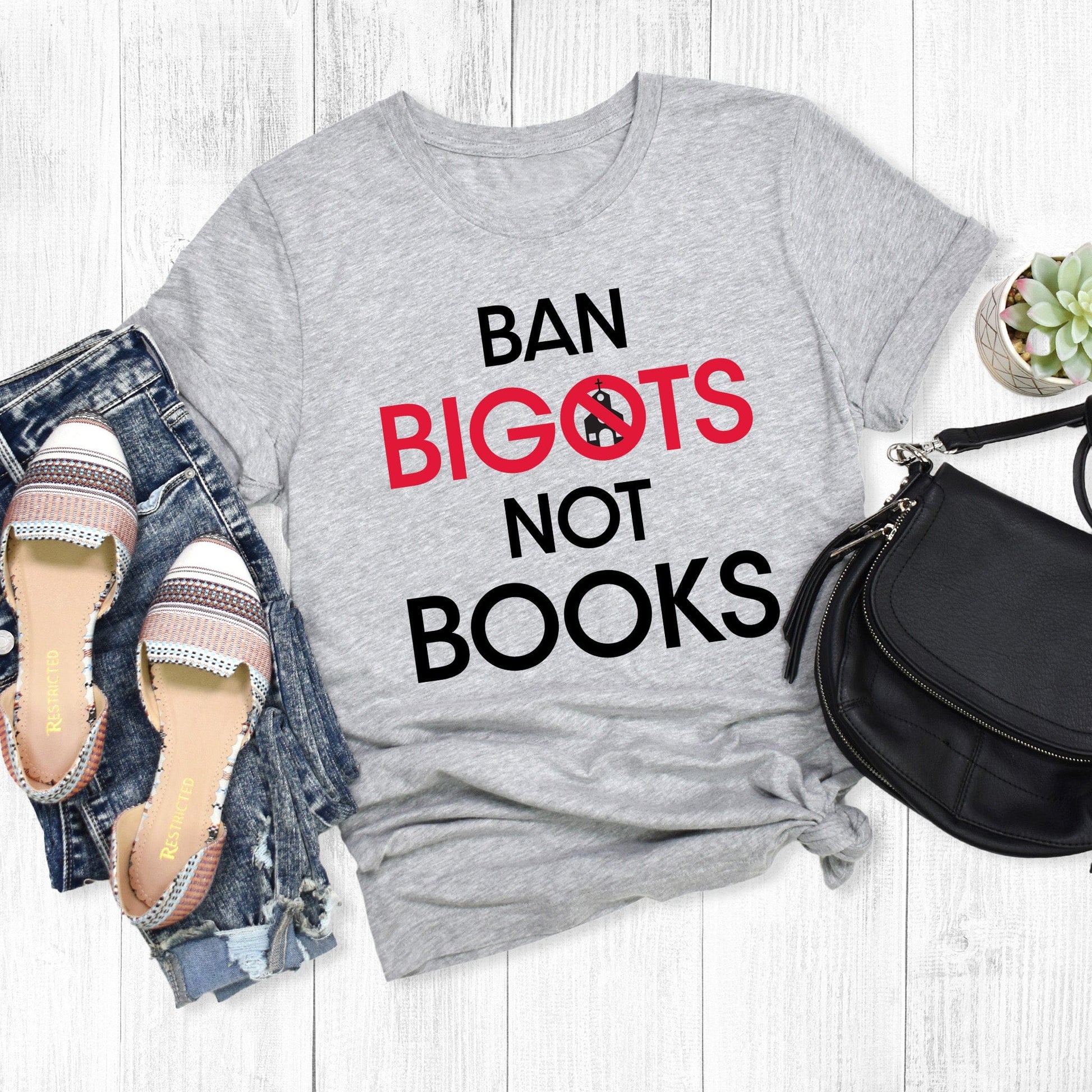 Athletic Heather color unisex t-shirt that says, “Ban Bigots Not Books” in all caps. Each word is on its own line. The word “Bigots” is red and the “O” has a line illustration of a church inside it with a slash to cross it out.