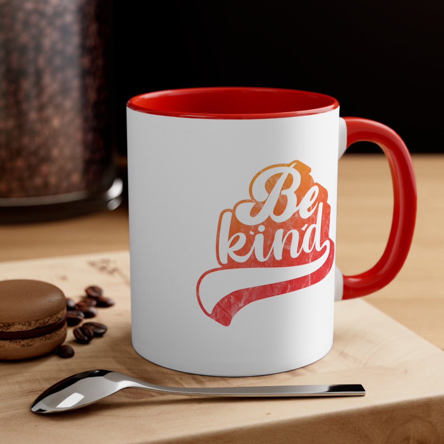 Contextual photo of an 11oz ceramic mug with a red handle and interior that says, “Be Kind” in a script font that is an orange gradient color. The “d” has a unique swoosh glyph extending below the word, adding a dynamic and flowing aesthetic to the text.