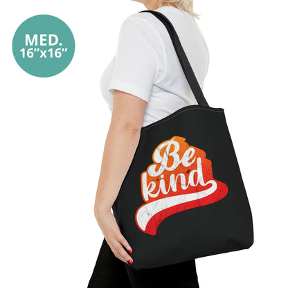 Contextual photo of a woman holding a medium 16x16” black tote bag that says, “Be Kind” in a script font that has an orange gradient. The “d” has a unique swoosh glyph extending below the word, adding a dynamic and flowing aesthetic to the text.