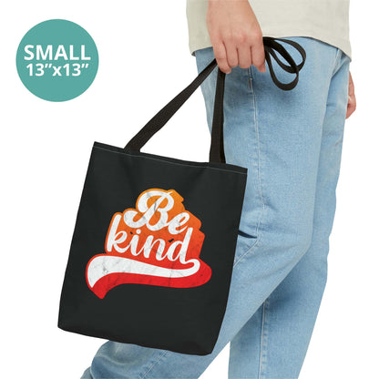 Contextual photo of a man holding a small 13x13” black tote bag that says, “Be Kind” in a script font that has an orange gradient. The “d” has a unique swoosh glyph extending below the word, adding a dynamic and flowing aesthetic to the text.