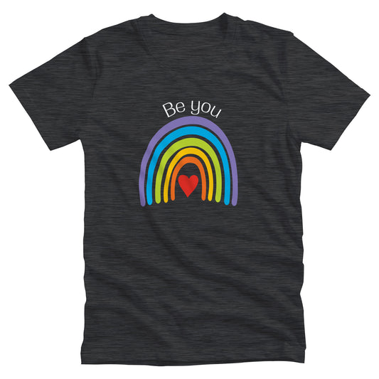 Dark Grey Heather color unisex t-shirt with an uneven illustration of a rainbow. The negative space inside the rainbow holds a red heart. There is text arched above the rainbow that says, “Be you.”