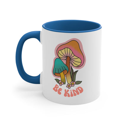 11oz ceramic mug with a blue handle and interior that says “Be Kind” on the front in a retro font with two retro mushrooms above the text. The text is slightly arched down.