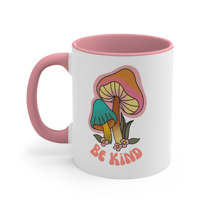 11oz ceramic mug with a pink handle and interior that says “Be Kind” on the front in a retro font with two retro mushrooms above the text. The text is slightly arched down.