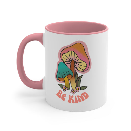 11oz ceramic mug with a pink handle and interior that says “Be Kind” on the front in a retro font with two retro mushrooms above the text. The text is slightly arched down.