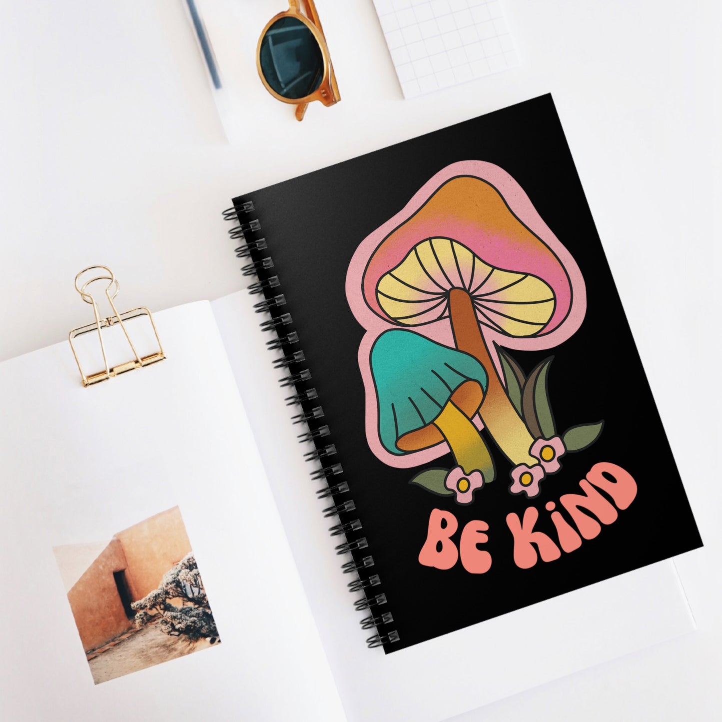 Contextual photo of a black spiral notebook that says “Be Kind” on the front in a retro font with two retro mushrooms above the text. The text is slightly arched down.