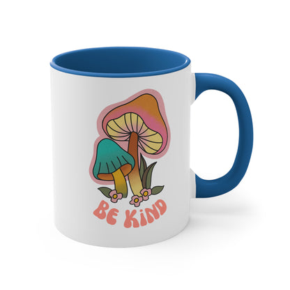 11oz ceramic mug with a blue handle and interior that says “Be Kind” on the front in a retro font with two retro mushrooms above the text. The text is slightly arched down.