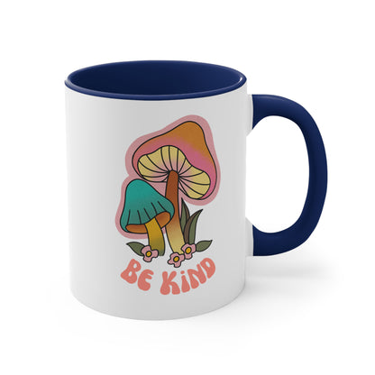 11oz ceramic mug with a navy blue handle and interior that says “Be Kind” on the front in a retro font with two retro mushrooms above the text. The text is slightly arched down.