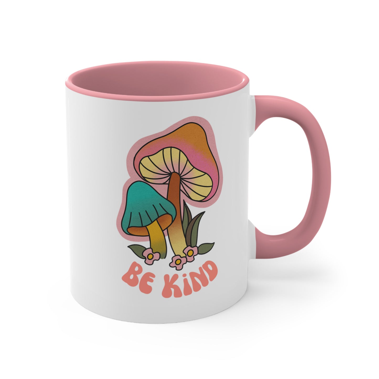 11oz ceramic mug with a pink handle and interior that says “Be Kind” on the front in a retro font with two retro mushrooms above the text. The text is slightly arched down.