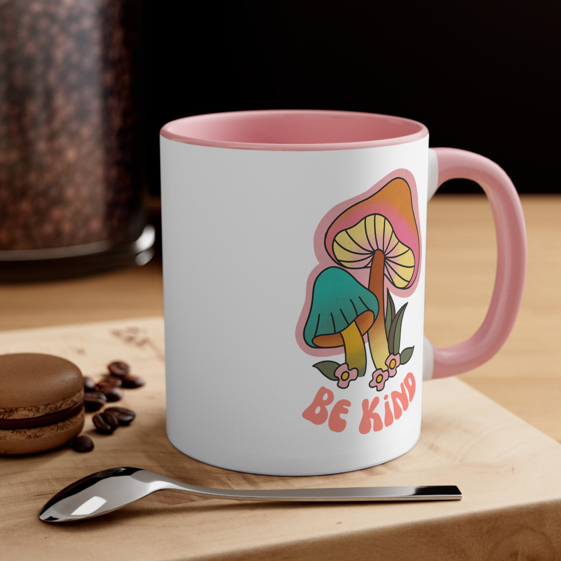 Contextual photo of an 11oz ceramic mug with a pink handle and interior that says “Be Kind” on the front in a retro font with two retro mushrooms above the text. The text is slightly arched down. The mug is on a wooden table with a spoon and a cookie beside it.