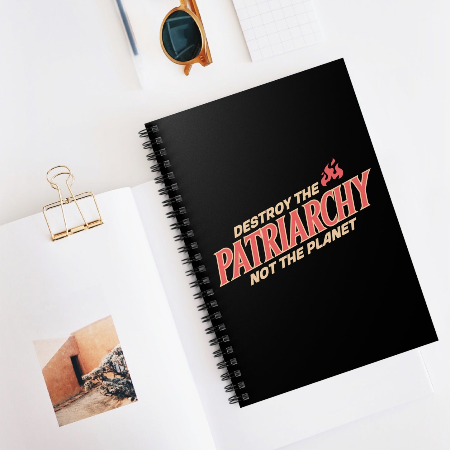 Contextual image of a black 6x8” ruled-line spiral notebook with text: 'DESTROY THE PATRIARCHY NOT THE PLANET slanted upwards. 'DESTROY THE' is on the first line with small flames after it, 'PATRIARCHY' is on the second line, and 'NOT THE PLANET' is on the third line.