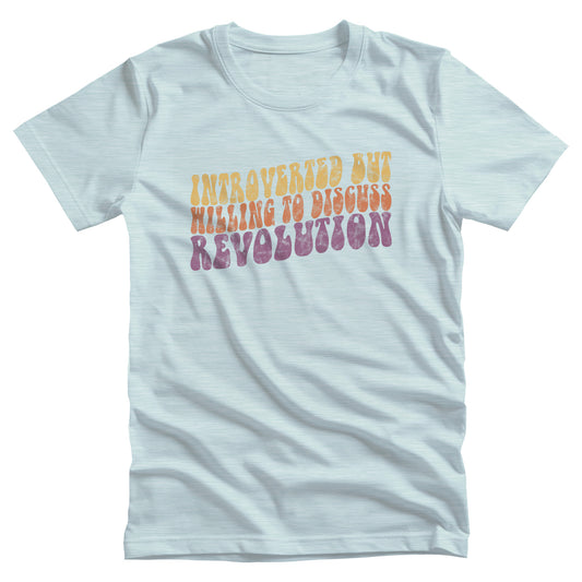 Heather Ice Blue color unisex t-shirt with a vintage graphic on it that looks worn and grungy. The graphic says “Introverted but willing to discuss revolution” in a retro font in all caps. Each line is a different color: “Introverted but” is a retro yellow, “willing to discuss” is a retro red, and “Revolution” is a retro purple. The graphic is a wavy shape and slopes upwards to the right.