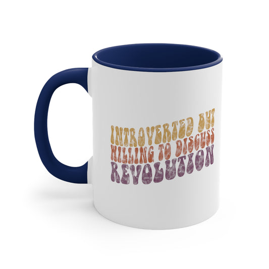 11oz white ceramic mug with a navy blue handle and interior. It has a vintage graphic on it that looks worn and grungy. The graphic says “Introverted but willing to discuss revolution” in a retro font in all caps. Each line is a different color: “Introverted but” is a retro yellow, “willing to discuss” is a retro red, and “Revolution” is a retro purple. The graphic is a wavy shape and slopes upwards to the right.