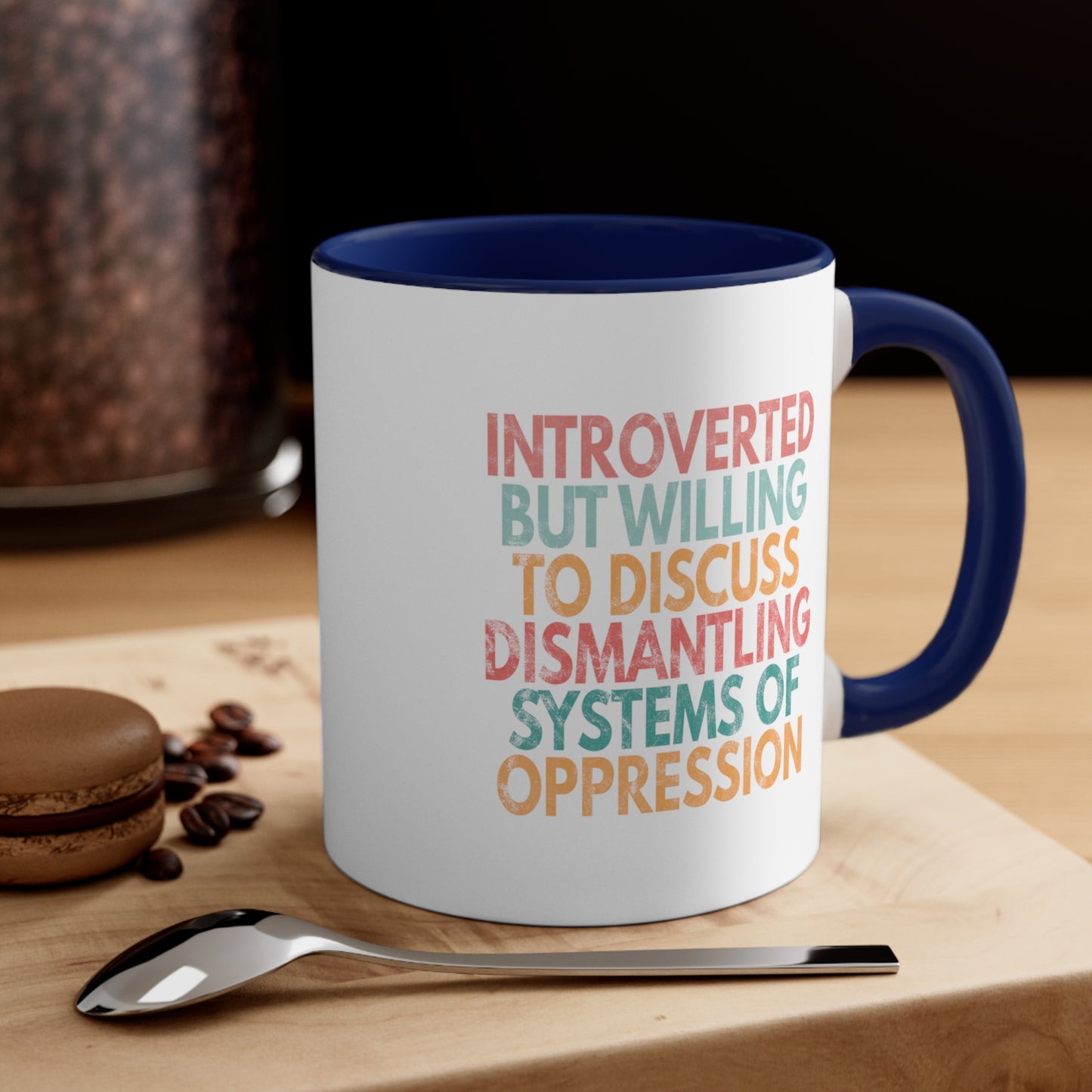 Context image of an 11oz ceramic mug with a navy blue interior and handle with a graphic that says “Introverted but willing to discuss dismantling systems of oppression.” The graphic is slightly distressed, and each line of the graphic is a different color. The mug is on a table with cookies and a spoon.