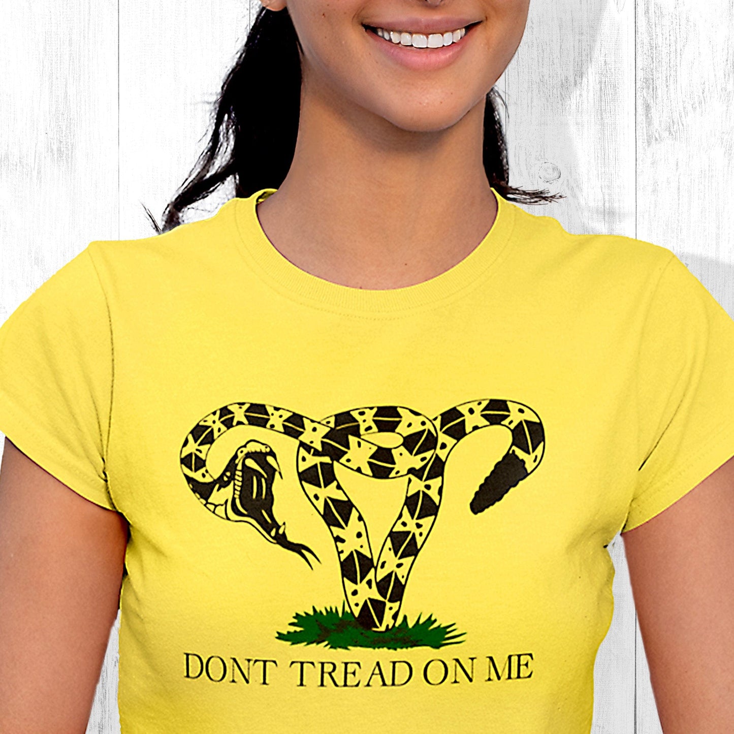 Yellow unisex t-shirt with an image of a rattlesnake coiled into the shape of a uterus. Its mouth is wide open ready to strike. Beneath the graphic, it says, “DONT TREAD ON ME” with no apostrophe in “DON’T” like the original.