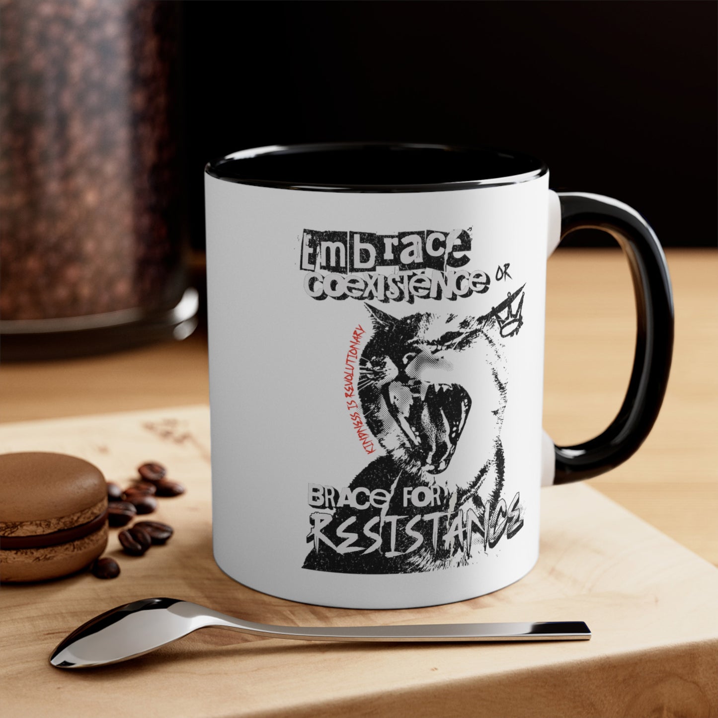 Contextual photo of an 11oz ceramic mug with a black interior and handle featuring the phrase 'Embrace Coexistence or Brace for Resistance' in a grunge font. The shirt includes an illustration of a cat meowing loudly with a small crown to the right, adding to the grunge aesthetic. Small red letters along the left side of the cat's face, written vertically say, ‘Kindness is revolutionary' in all caps. The mug is on a table with a spoon and a cookie.