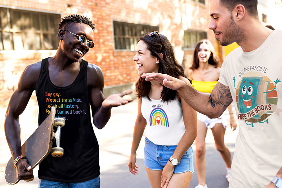 Three diverse friends wearing equality shirts. The man on the left is wearing a black tank top that say, "Say gay. Protect trans kids. Teach all history. Read banned books. Show love." in rainbow colors. The woman in the middle is wearing a tank that says, "Be you" arched over a graphic of a rainbow. The man on the right is wearing a t-shirt that says, "BAN THE FASCISTS" arched over a circular graphic of a cartoon book with a happy face leaping for joy. Over the book is a ribbon that says, "FREE THE BOOKS."