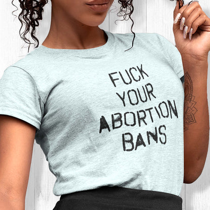 Heather Ice Blue color unisex t-shirt that says, “FUCK YOUR ABORTION BANS” in a stencil font with ink bleeding slightly outside of the text.