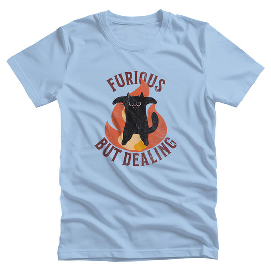 Baby Blue color unisex t-shirt that says “Furious but dealing” in all caps. The word “furious” is arched over an image of an angry black cat standing up on its hind legs with its arms spread with a large flame behind it. “But Dealing” is arched the opposite way underneath the graphic. The graphic is retro inspired with a worn look.