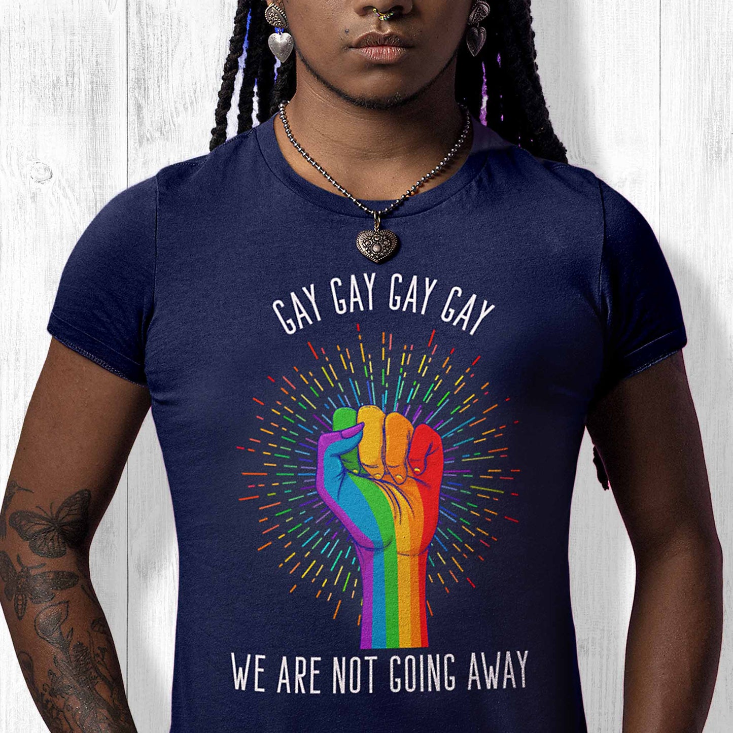 Navy Blue color unisex t-shirt that says, “Gay Gay Gay Gay We Are Not Going Away” with a graphic of a rainbow fist with a thin, rainbow-colored starburst behind it. Arched over the top of the starburst are the words “Gay Gay Gay Gay” in all caps. The rest of the text is beneath the graphic on one horizontal line.