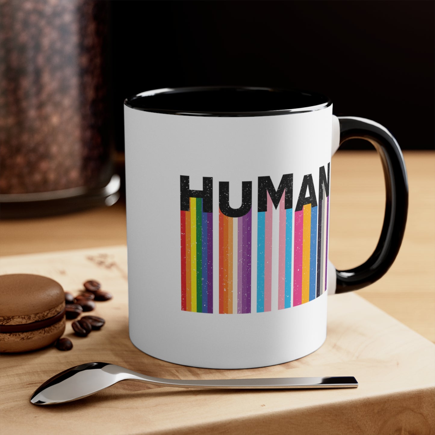 Contextual photo featuring an 11oz ceramic mug with a pink interior and handle. It has a colorful and dynamic image featuring the word "HUMAN" written in an artistic style.  Each letter is arranged to create a wavy pattern. There are stripes of colors coming down from the bottom of each letter representing different sexualities in the LGBTQ+ community. The "H" represents Gay, the "U" represents Lesbian, the "M" represents Trans, the "A" represents Pansexual, and the "N" represents Asexual.