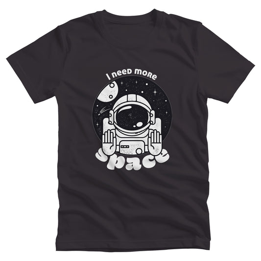 Dark Grey unisex t-shirt that says, “I need more space”. The text is a retro font. There is a graphic of an astronaut inside a circle with their hands in the stop position. “I need more” is arched over the graphic, and “space” is reverse-arched over the bottom of the graphic.