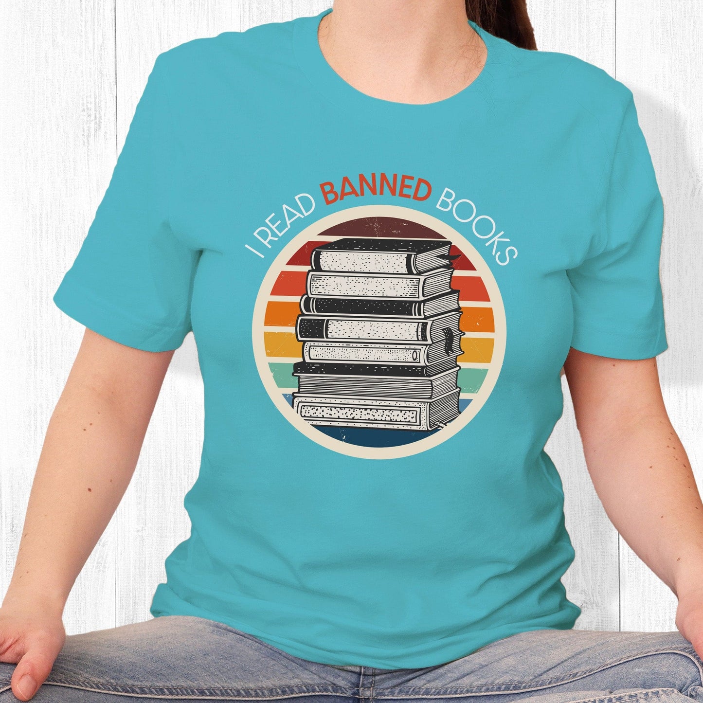 Turquoise unisex t-shirt with a circular graphic of stacked old books with a vintage sunset made from horizontal lines behind it. “I READ BANNED BOOKS” is arched over the graphic with the word. “BANNED” an orange color.