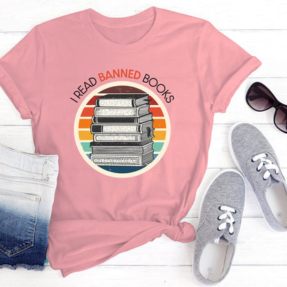 Pink unisex t-shirt with a circular graphic of stacked old books with a vintage sunset made from horizontal lines behind it. “I READ BANNED BOOKS” is arched over the graphic with the word. “BANNED” an orange color.