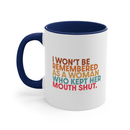 11oz ceramic mug with a navy blue handle and interior that says, “I won’t be remembered as a woman who kept her mouth shut” in all caps. The phrase is on 5 lines, with each line being a different color. “I won’t be” is brown, “remembered” is orange, “as a woman” is yellow-orange, “who kept her” is blue-green, and “mouth shut” is pink-red. 