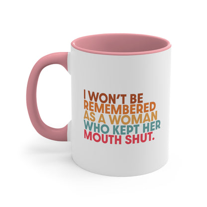 11oz ceramic mug with a pink handle and interior that says, “I won’t be remembered as a woman who kept her mouth shut” in all caps. The phrase is on 5 lines, with each line being a different color. “I won’t be” is brown, “remembered” is orange, “as a woman” is yellow-orange, “who kept her” is blue-green, and “mouth shut” is pink-red. 