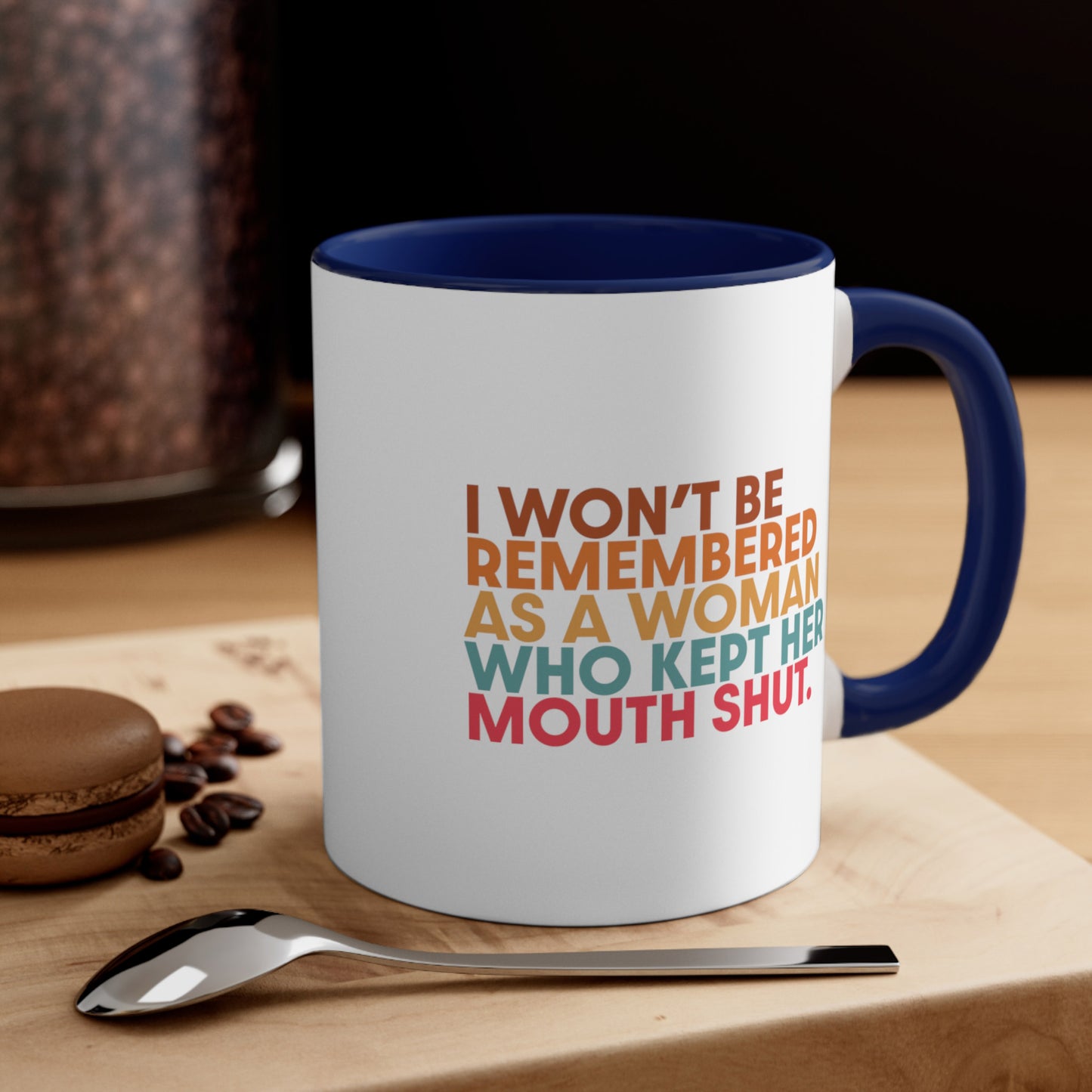 Contextual photo of an 11oz ceramic mug with a navy blue handle and interior that says, “I won’t be remembered as a woman who kept her mouth shut” in all caps. The phrase is on 5 lines, with each line being a different color. “I won’t be” is brown, “remembered” is orange, “as a woman” is yellow-orange, “who kept her” is blue-green, and “mouth shut” is pink-red. 