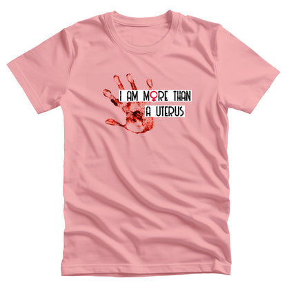 Pink unisex t-shirt that says, “I Am More Than a Uterus” in all caps. “I Am More Than” is on one line and “a Uterus” is on a separate line. The text all has a thin white rectangle behind it that expands the length of the text. To the left is a red handprint.