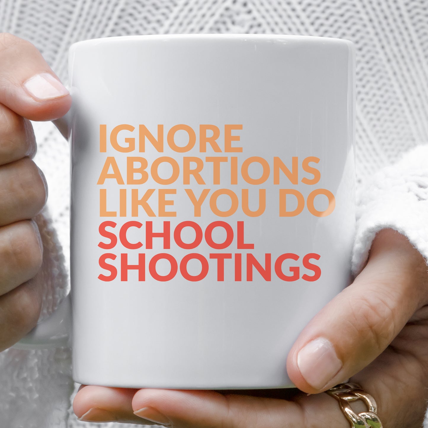 A woman holding a white 11oz ceramic mug that says “Ignore abortions like you do school shootings” in all caps in an orange font. The words “school shootings” are in red. The handle is on the left side in this image.