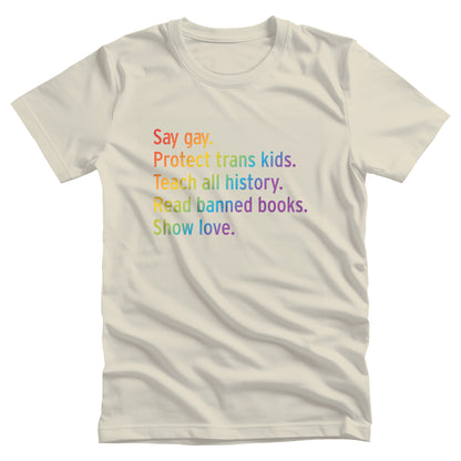 Natural color unisex tank top with text that says “Say gay. Protect Trans Kids. Teach all history. Read banned books. Show love.” Each sentence is on its own line, and the entire graphic is colored with a rainbow gradient.