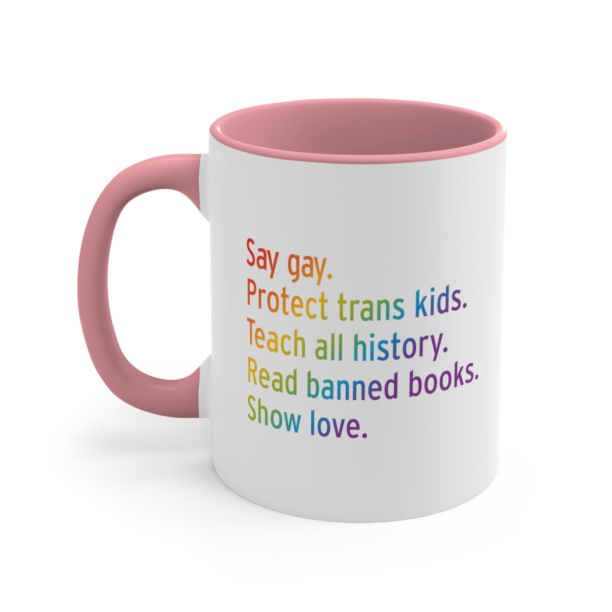 11oz white ceramic mug colored pink on the inside of the mug and the handle. The text says “Say gay. Protect Trans Kids. Teach all history. Read banned books. Show love.” Each sentence is on its own line, and the entire graphic is colored with a rainbow gradient.