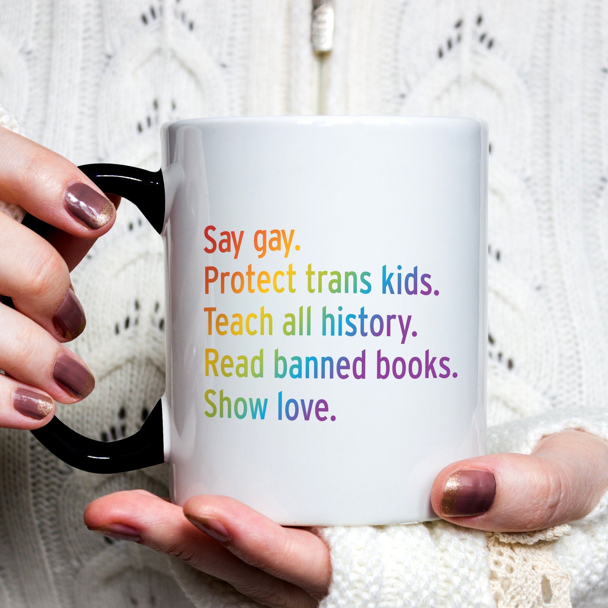 A woman wearing a white sweater holding an 11oz white ceramic mug colored black on the inside of the mug (which can’t be seen at this angle) and the handle. The text says “Say gay. Protect Trans Kids. Teach all history. Read banned books. Show love.” Each sentence is on its own line, and the entire graphic is colored with a rainbow gradient.