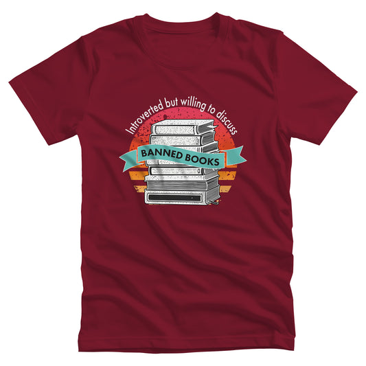 Cardinal color unisex t-shirt with a graphic that says, “Introverted but willing to discuss banned books.” The image has stacked books with a circular retro sunset behind it. “Introverted but willing to discuss” is arched over the graphic, and “banned books” is inside a wavey blue-green colored ribbon.