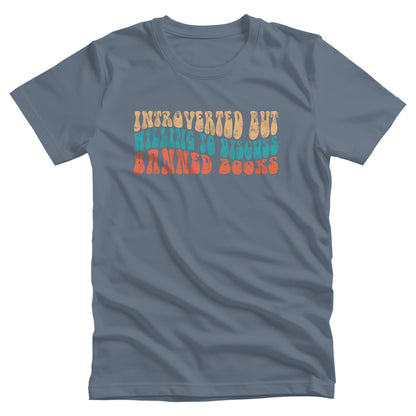 Steel Blue color unisex t-shirt with a graphic that says “Introverted but willing to discuss banned books” in a retro, 1970s bubble font. Each line is a different color. The graphic is slightly distressed.